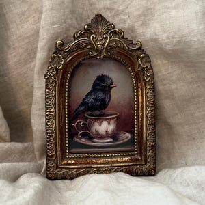 May include: A black bird perched on a vintage porcelain teacup with a floral design. The bird is sitting on a saucer and the image is framed in a gold ornate frame.