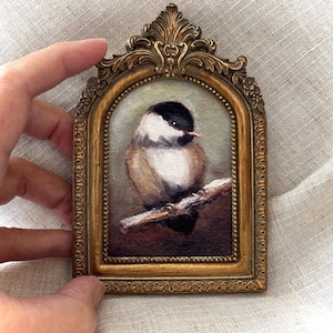 May include: A small, framed painting of a bird perched on a branch. The bird has a black head, white cheeks, and a brown body. The ornate gold frame has a decorative top and a curved shape.