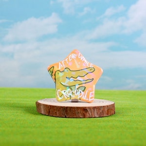 May include: A star-shaped button with a light orange background and a cartoon crocodile with the text "After While Crocodile" on it.