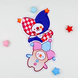 May include: A whimsical sticker featuring two cartoon clowns stacked on top of each other. The top clown wears a blue hat and a red and white checkered collar. The bottom clown is pink and blue. Several star-shaped decorations are scattered around the clowns.