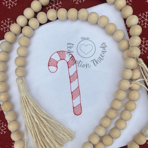 May include: A white square fabric with a red and white candy cane embroidery design. The fabric is surrounded by a wooden bead garland with tassels. The background is red with white snowflake patterns. The text "Redemption Threads" is also visible.