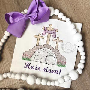 May include: White fabric with an embroidered image of three crosses and a stone tomb with the text "He is risen!"  The image is surrounded by a purple bow and a string of white beads.