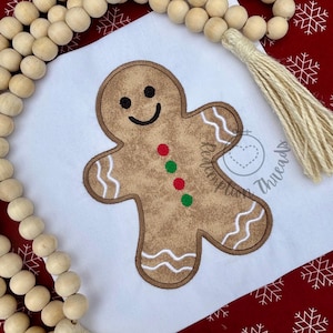 May include: A brown gingerbread man applique with a smiling face and red and green buttons is centered on a white background. The applique is outlined in a darker brown. White wavy lines are on the arms and legs. A wooden bead garland and tassel are in the image.