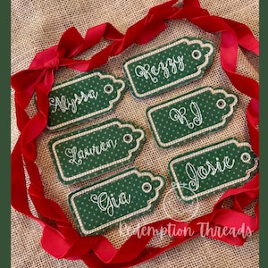 Puede incluir: Seis etiquetas de regalo verdes con lunares blancos y nombres bordados en blanco: Alyssa, Peggy, RJ, Lauren, Gia y Josie. Las etiquetas están dispuestas en círculo sobre un fondo de arpillera con una cinta roja.
