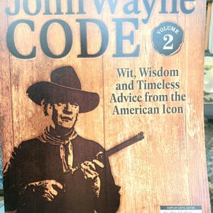 Puede incluir: Portada del libro The John Wayne Code, Volumen 2. Imagen en sepia de John Wayne con sombrero de vaquero, sosteniendo un rifle. El texto incluye "Wit, Wisdom and Timeless Advice from the American Icon."