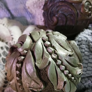 May include: Two decorative, spherical objects with ornate, leaf-like designs. The front object is silver-toned with detailed leaves and a textured base. The second object is bronze-coloured with a swirling pattern.