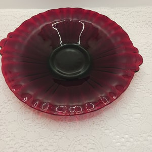 Könnte beinhalten: Eine rubinrote Glasservierschale mit gerüschtem Rand und schwarzem Mittelteil. Die Schale hat eine runde Form mit leicht erhöhtem Mittelteil und zwei kleinen Griffen. Das Glas hat eine glänzende Oberfläche.