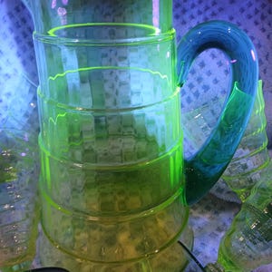 May include: A translucent neon yellow glass pitcher featuring a tiered, ribbed cylindrical body and a contrasting solid teal blue curved handle. The glass exhibits a bright glow, and the design includes a small pouring spout at the top rim.