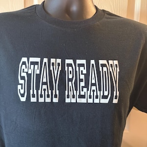 May include: A black short-sleeved t-shirt featuring the text STAY READY printed in a bold, white, block-style font across the chest. The garment has a classic crew neckline and is displayed on a mannequin to show the fit and design clearly.