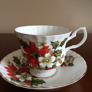May include: A white teacup and saucer with a floral design featuring red poinsettia flowers, white flowers, and green holly leaves.
