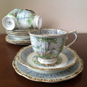 May include: A collection of vintage teacups and saucers. The cups and saucers feature a light blue and white design with gold edging. The teacups display a landscape scene with trees and flowers. The set includes several cups and saucers.