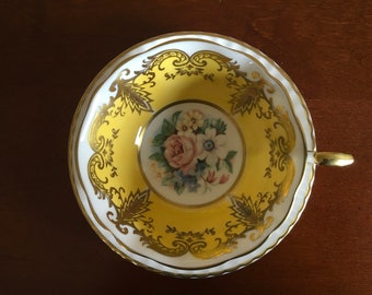 Può includere: Una tazza da tè bianca con un motivo floreale giallo e oro. La tazza ha un bordo ondulato e un manico dorato.