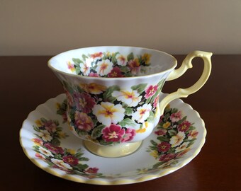 May include: A white teacup and saucer with a floral pattern. The cup and saucer are decorated with yellow, pink, and white flowers on a black background. The saucer has a scalloped edge.