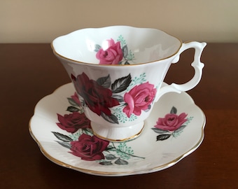 May include: A white teacup and saucer set with a floral pattern featuring pink and red roses. The set has a gold trim around the edges.