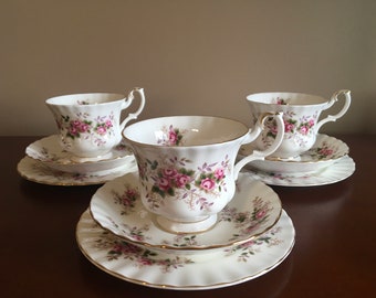 May include: Three white teacups and saucers with a floral pattern of pink roses and green leaves. The cups and saucers have a gold trim.