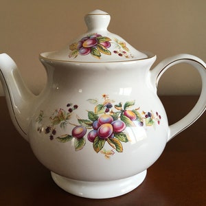 Peut inclure: Une théière en céramique blanche avec un motif floral représentant des prunes violettes et des feuilles vertes. La théière est ornée d'une bordure dorée autour du couvercle et du bec.