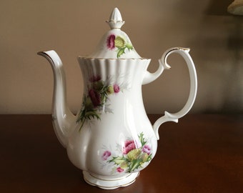 May include: White porcelain teapot with a gold trim and a floral design featuring purple and green thistles.