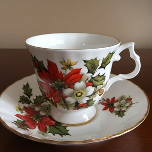 May include: A white teacup and saucer with a floral design featuring red poinsettia flowers, white flowers, and green holly leaves. The cup and saucer have a gold trim.