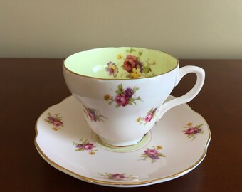 May include: A white teacup and saucer set with a floral pattern. The cup has a pale green interior and a gold rim. The saucer has a scalloped edge and a gold rim.