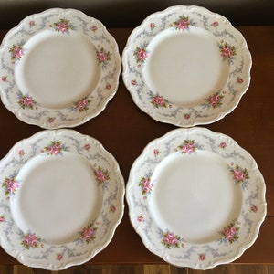 Op de afbeelding: Set van vier witte keramische dinerborden met een delicaat bloemenontwerp met roze rozen en blauwe accenten. De borden hebben een gegolfde rand en een gouden rand.