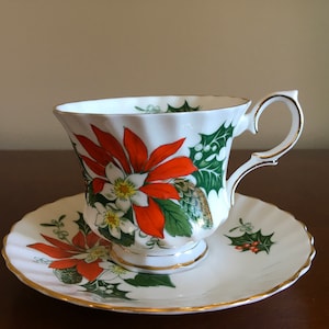 May include: A white teacup and saucer with a gold rim. The cup and saucer are decorated with a red and green poinsettia floral pattern.