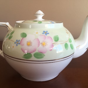 May include: A white teapot with a green and white floral design. The teapot has a gold trim around the rim and a gold handle.