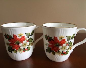 May include: Two white ceramic mugs with gold rims and a Christmas floral design featuring red poinsettia flowers, white flowers, green holly leaves, and red berries.