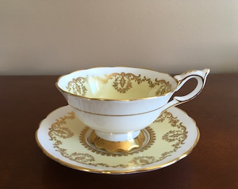 May include: A white teacup and saucer with a yellow and gold floral pattern. The cup has a handle and the saucer has a scalloped edge.