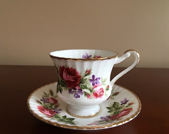 May include: A white teacup and saucer with a floral pattern featuring pink, red, and purple roses and leaves. The cup and saucer have a gold trim.