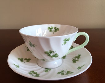 May include: A white teacup and saucer set with a green shamrock pattern. The cup has a light green handle.