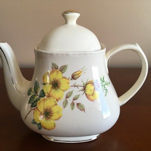 May include: A white ceramic teapot with a floral design featuring yellow roses. The teapot has a lid and a handle.