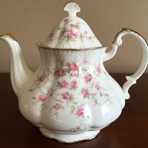 Peut inclure: Une théière blanche avec un motif floral de roses roses et de feuilles vertes. La théière a une bordure dorée autour du couvercle et de la base.
