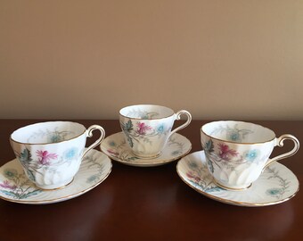 May include: Three white porcelain teacups and matching saucers featuring a delicate floral pattern in shades of pink, blue, and grey. Each piece has a fluted texture and is finished with a thin gold trim along the scalloped edges.