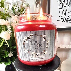 May include: A red three-wick candle with a farmhouse-themed label featuring a wooden background with white cotton bolls and the text "FARM" in a black and white font. The label also includes the text "MIDWEST URBAN FARMHOUSE". The candle is sitting on a black pedestal with a black and white checkered tablecloth underneath.
