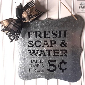 May include: A rustic metal sign with a distressed finish, featuring the text "FRESH SOAP & WATER HAND TOWELS FREE 5¢". The sign is decorated with a black and white plaid bow.