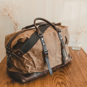 May include: A brown canvas and leather duffel bag with a long shoulder strap and multiple pockets. The bag has a zipper closure and a buckle on the front.