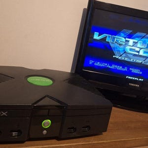May include: Black Xbox console with a green power button and logo, beside a flat-screen TV showing "Virtua Cop 3". The console is on a wooden surface.