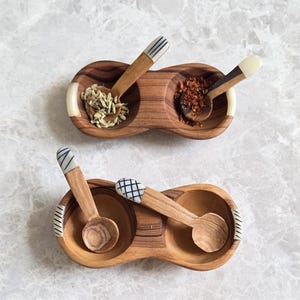 Peut inclure: Trois plats à épices en bois avec cuillères attachées. Chaque plat comporte deux compartiments remplis d'épices différentes. Les cuillères ont des manches décoratifs à rayures ou à carreaux blancs et noirs. Les plats sont en bois brun clair.