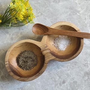 May include: Wooden salt and pepper serving dish with two bowls and a small spoon. One bowl contains black pepper, the other salt. A small bouquet of yellow flowers is in the background. The dish is on a grey marble surface.