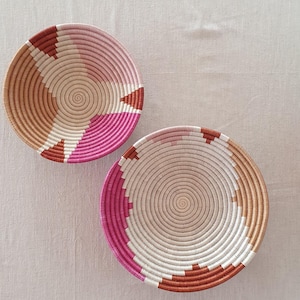 Pode incluir: Duas cestas de vime com um padrão geométrico rosa, branco e castanho. As cestas são redondas e têm uma ligeira curvatura nas laterais.