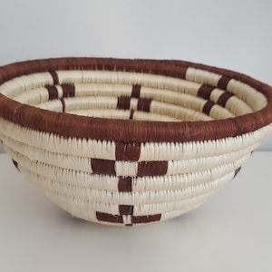 Pode incluir: Uma cesta redonda tecida à mão. Apresenta uma base creme com padrões geométricos castanhos e uma borda castanha. A cesta é feita de fibras naturais e é adequada para armazenamento ou fins decorativos.