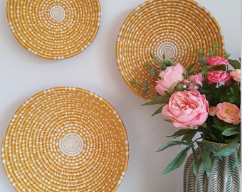 May include: Three round woven wall baskets in a yellow and white pattern. The baskets are hanging on a white wall. A vase with pink flowers sits on a white surface in front of the baskets.