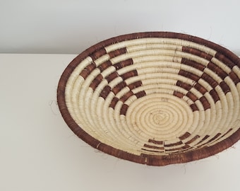 May include: A large, round woven basket with a brown and white pattern. The basket is made of natural materials and has a wide, shallow opening.