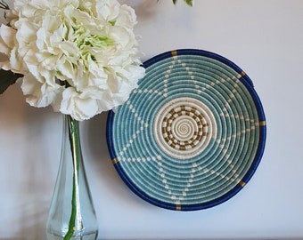 Peut inclure: Un panier mural tissé bleu et blanc avec un accent doré. Le panier est rond et présente un motif circulaire. Il est suspendu à un mur blanc avec un arrangement de fleurs blanches dans un vase en verre transparent au premier plan.