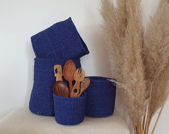 May include: Four blue woven baskets of varying sizes, stacked and arranged on a light brown surface. The baskets are filled with wooden utensils, including a spoon, a fork, and a wooden carving.