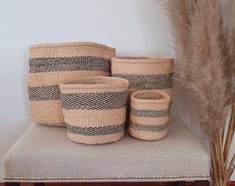 May include: Four woven storage baskets of different sizes, with a beige and black striped design. The baskets are displayed on a light-coloured upholstered bench. The background features a white wall and dried ornamental grass in a glass vase.