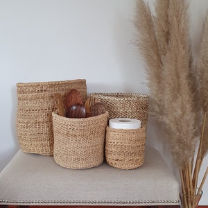 Könnte beinhalten: Vier geflochtene Körbe in verschiedenen Größen, aus natürlichen Materialien, sind auf einer hellbeigen Oberfläche angeordnet. Die Körbe sind leer, bis auf den kleinsten, der eine Rolle weißes Papiertuch enthält.