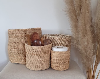 May include: Four woven baskets in various sizes, made of natural materials, are arranged on a light beige surface. The baskets are empty except for the smallest one, which contains a roll of white paper towels.
