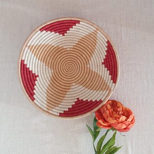 May include: A woven basket with a red and beige geometric pattern. The basket is round and has a flat bottom. It is sitting on a white fabric surface.