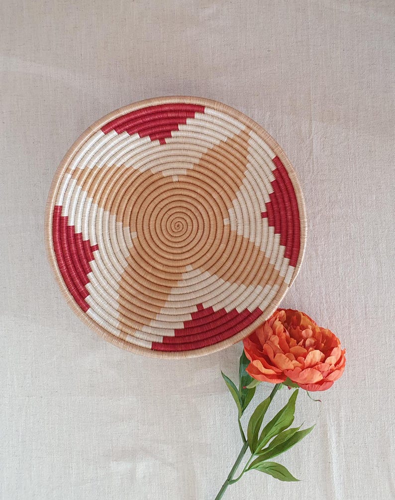 May include: A woven basket with a red and beige geometric pattern. The basket is round and has a flat bottom. It is sitting on a white fabric surface.